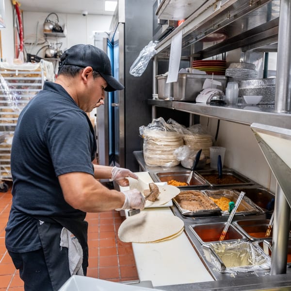 A chef in a black cap and apron prepares a burrito with meat and other fillings.
