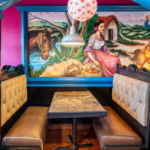 A vibrant Mexican mural with a woman, horse, and landscape behind a restaurant booth with a marble table.