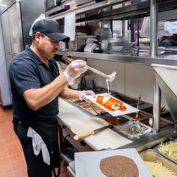 A chef in a black cap and gloves drizzles white sauce over a red-sauced burrito in a kitchen.